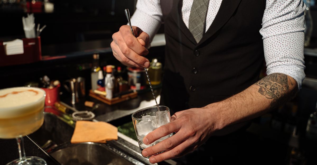 Bartender with tattooed arm mixing cocktail with ice cubes in a classy nightclub.